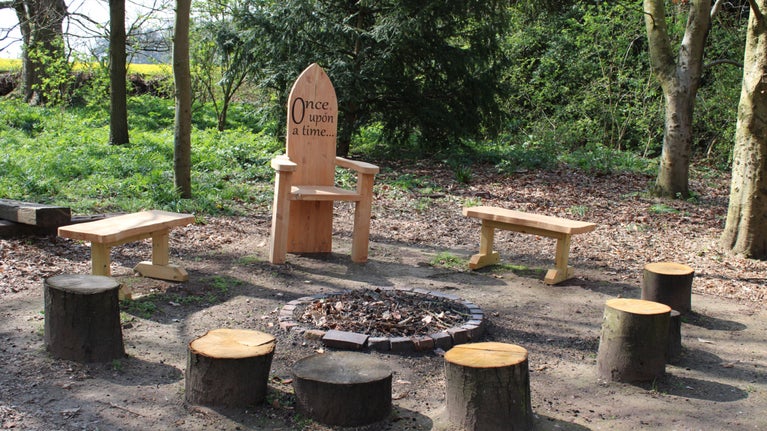 The new wooden storytelling chair, with 'Once upon a time' carved on the back rest is next to the firepit, surrounded by smaller log seats and wooden benches.